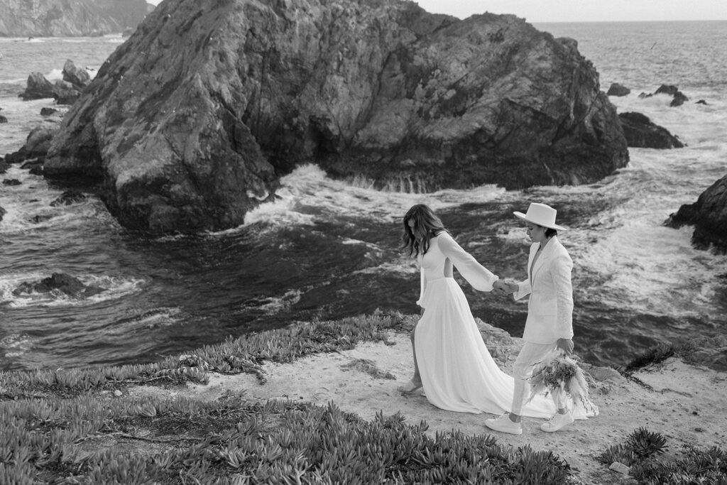 couple holding hands and walking in front of california cliffs and ocean